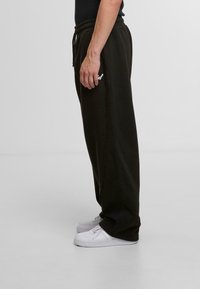 Dark green sweatpants with a relaxed fit, elastic waistband, and drawstring. Accented with a white logo on the thigh. Worn with white sneakers.