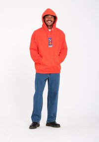 Oranje hoodie met een voorzak en een grafische print. Gecombineerd met blauwe jeans en zwarte schoenen. Gladde textuur, casual pasvorm.