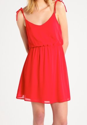 Žena v červených šatech bez rukávů s tenkými ramínky uvázanými na ramenou, zvýrazněným pasem a volánovou sukní na bílém pozadí.