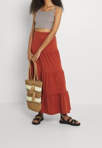 Šedý zkrácený tank top, vrstvená maxi sukně v cihlové barvě, černé sandály a tkaná taška s hnědými a krémovými pruhy a třásněmi.