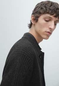 Dunkle, strukturierte Strickjacke mit erhabenem Rippenmuster, ausgestattet mit einem knöpfbaren Kragen und dezenten Details. Trägt man über einem schwarzen Hemd.