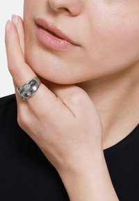 Anello d'argento con una superficie liscia e lucida, indossato su un dito, abbinato a una mano leggermente appoggiata sul viso. La pelle è di tonalità chiara.