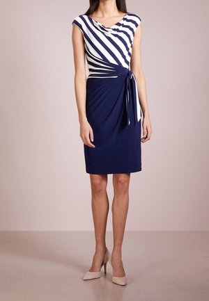 Femme portant une robe marine longueur genoux avec un haut à rayures diagonales blanches et marines et une ceinture latérale, associée à des talons hauts beige.