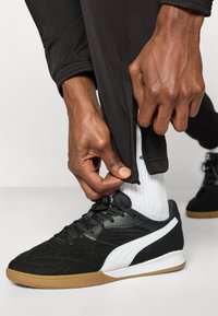 Μαύρα αθλητικά παπούτσια με υφασμάτινη υφή, λευκές λεπτομέρειες και λαστιχένιες σόλες. Το μοντέλο φοράει μαύρο παντελόνι με φερμουάρ.