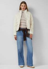 Kremowa pikowana kurtka z wysokim kołnierzem, w zestawieniu z brązowym swetrem związanym w pasie, pasiastym golfem i szerokimi niebieskimi dżinsami.