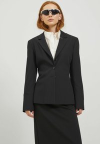 Blazer nero sartoriale con maniche lunghe e revers a punta, abbinato a una camicia bianca con colletto e una gonna nera coordinata. Occhiali da sole neri indossati.