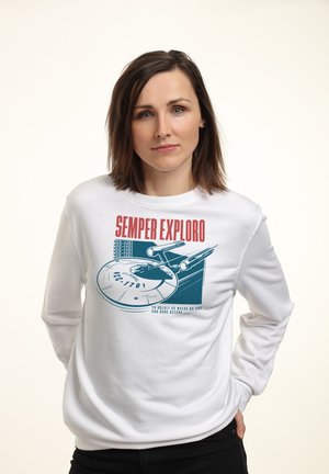 Femme portant un sweat-shirt blanc avec un texte rouge "Semper Exploro" et un vaisseau spatial graphique bleu avec le texte "NCC-1701" à l'avant, debout les mains dans les poches.