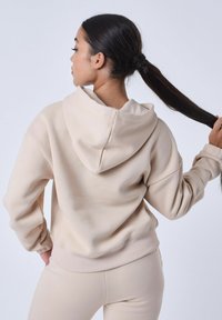 Beige hoodie met een relaxte pasvorm, voorzak, trekkoord bij de capuchon en geribbelde manchetten. Gecombineerd met bijpassende beige leggings. Zachte textuur.