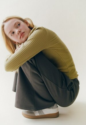 Jeune femme aux cheveux blonds, portant un haut vert côtelé, un pantalon large foncé et des baskets blanches, assise accroupie et regardant sur le côté.