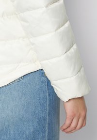 Puchowa biała kurtka z pikowanym wzorem, o lekko błyszczącej fakturze i dopasowanych rękawach. Połączona z niebieską spódnicą dżinsową.