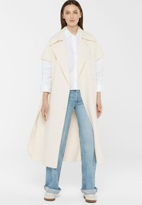 Frau in einem langen cremefarbenen Mantel mit breiten Revers über einem weißen Hemd, hellblauen, hochgekrempelten Jeans und weißen Sneakers vor einem weißen Hintergrund.
