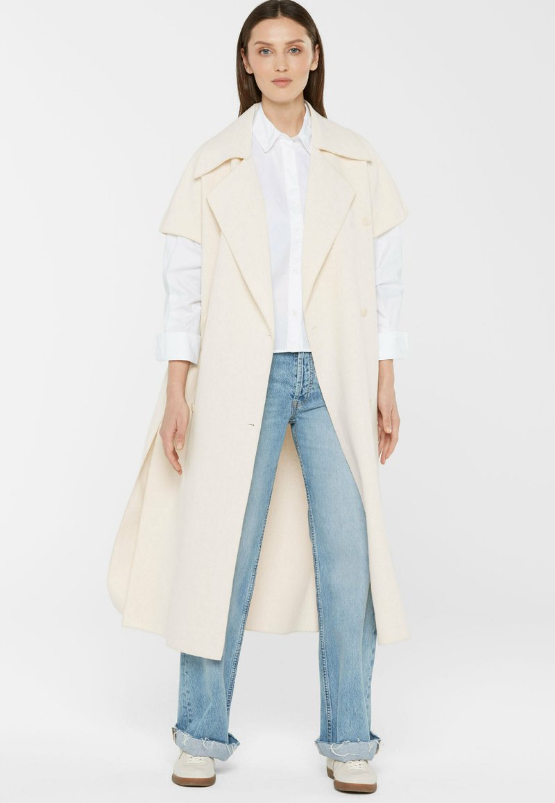 Frau in einem langen cremefarbenen Mantel mit breiten Revers über einem weißen Hemd, hellblauen, hochgekrempelten Jeans und weißen Sneakers vor einem weißen Hintergrund.