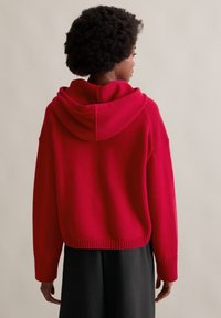 Maglione con cappuccio rosso realizzato in morbido tessuto a maglia, con vestibilità comoda, polsini e orlo a costine. Indossato sopra una gonna nera, con design corto.