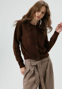 Braune Wollstrickjacke mit rundem Ausschnitt, Knopfleiste vorne und gerippten Bündchen, kombiniert mit beigen, hoch taillierten Hosen mit Bindegürtel.