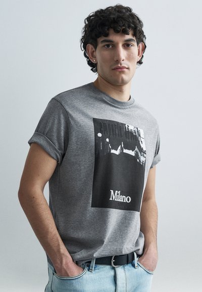 T-shirt gris en coton à manches courtes avec une impression graphique en noir et blanc, texte "Milano" en bas et manches retroussées.