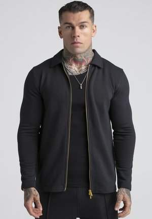 Mann mit Hals- und Handtattoos, trägt eine schwarze Jacke mit Reißverschluss und ein schwarzes Shirt, schaut nach vorne vor schlichtem Hintergrund.