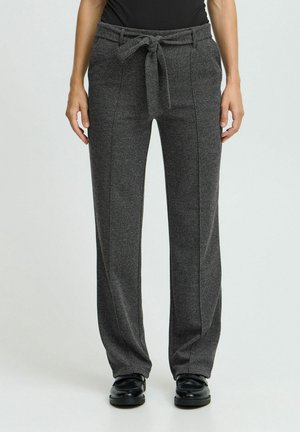 Frau trägt graue Hose mit hoher Taille und vorn gebundenem Gürtel, kombiniert mit schwarzen Schuhen und schwarzem Oberteil, stehend vor einfachem Hintergrund.