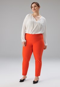 Weiße Bluse mit langen Ärmeln, V-Ausschnitt und Kordelzug, kombiniert mit enger oranger Hose und schwarzen, mit Schleifen verzierten Absatzschuhen. Schlichter Hintergrund.