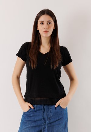 Giovane donna con capelli castani lunghi che indossa una t-shirt nera e jeans blu, in piedi con le mani nelle tasche contro uno sfondo neutro.