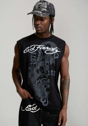 Man met gevlochten haar die een zwart mouwloos Ed Hardy-shirt met drakenprint draagt en een donkere pet met vleugelige schedelontwerp, terwijl hij naar beneden kijkt.