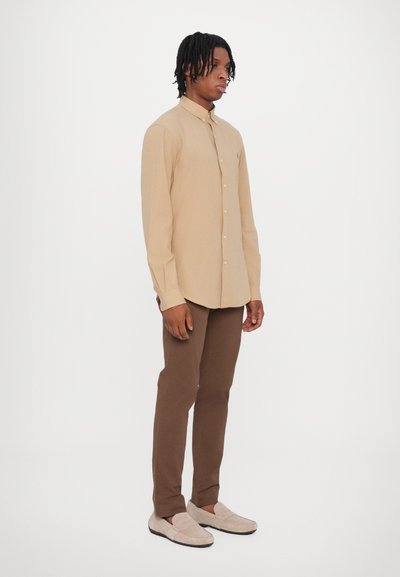 Camisa bege com botões, de mangas longas, combinada com calças ajustadas castanhas e mocassins bege. Tecido suave, design minimalista, detalhe de logo discreto.