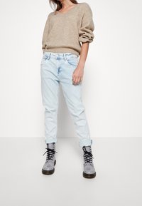 Frau in beigem Strickpullover, hellblauen, hochgekrempelten Jeans und glänzenden grauen Schnürstiefeln, die vor einem weißen Hintergrund steht.