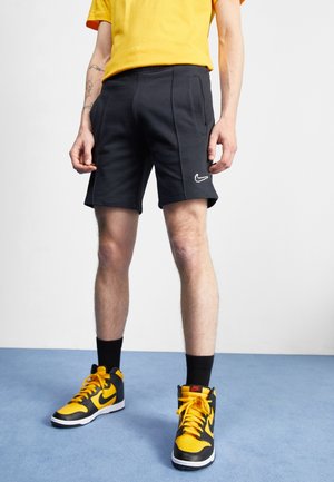 Mężczyzna w czarnych szortach Nike, żółto-czarnych wysokich butach Nike, czarnych skarpetkach i żółtej koszulce, stojący na niebieskiej podłodze.