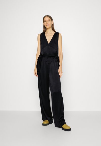 Top senza maniche in satin nero con scollatura a V, abbinato a pantaloni neri a gamba larga con vita regolabile e scarpe gialle a contrasto.