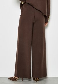 Braune, weit geschnittene Strickhosen mit glatter Textur und elastischem Bund, kombiniert mit spitzen braunen High Heels. Goldene Knopfakzente an der Oberseite.