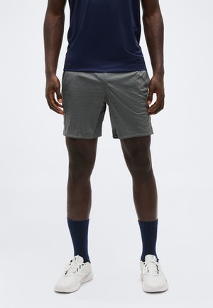 Moški v temnomodri majici, sivih športnih kratkih hlačah, temnomodrih nogavicah in belih supergah stoji ob belem ozadju.