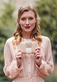 Béžová kožená peněženka s uzávěrem na knoflíček a vyraženým logem, držena vztyčená proti měkkým růžovým šatům s plisovanou texturou a knoflíky.