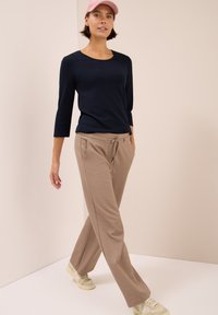 Marineblaues Langarmshirt mit Rundhalsausschnitt, kombiniert mit beigen Sweatpants mit Kordelzug. Das Modell trägt helle Sneakers und eine pinkfarbene Kappe.