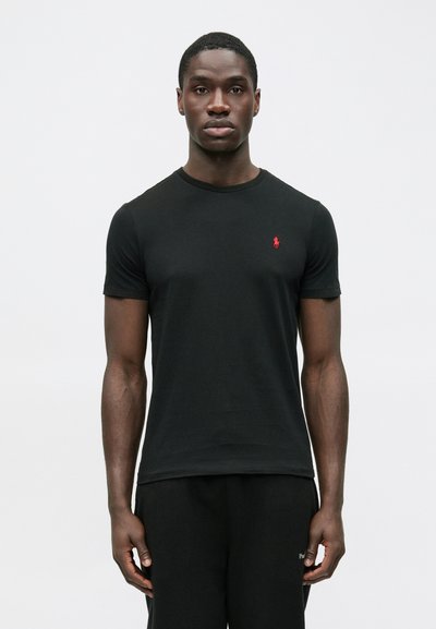 Młody mężczyzna w dopasowanej czarnej koszulce z czerwonym logo na piersi, stojący prosto na tle jednolitego, jasnoszarego tła.