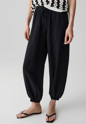 Pantaloni neri in lino dal taglio ampio con vita elastica e polsini, indossati con sandali infradito neri e top nero e bianco all'uncinetto.