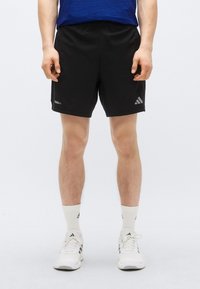 Čierne športové šortky s elastickým pásom a jemnými logovými detailmi v sivej farbe. Textúrovaná látka, doplnená bielymi športovými ponožkami a topánkami.