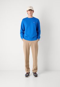 Blauwe sweatshirt met ronde hals, beige broek en veelkleurige sneakers. Pet met zwart "GANT" logo. Gladde stofstructuren zichtbaar.