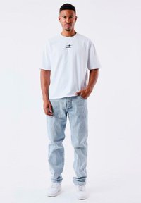 Camiseta blanca de manga corta con un pequeño gráfico negro en el pecho, combinada con unos vaqueros de denim azul claro y zapatillas blancas.