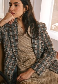 Blazer in tweed con motivo a quadretti scuri e chiari, abbinato a un abito dorato dalla texture ricca. Entrambi presentano dettagli intricati e forme strutturate.