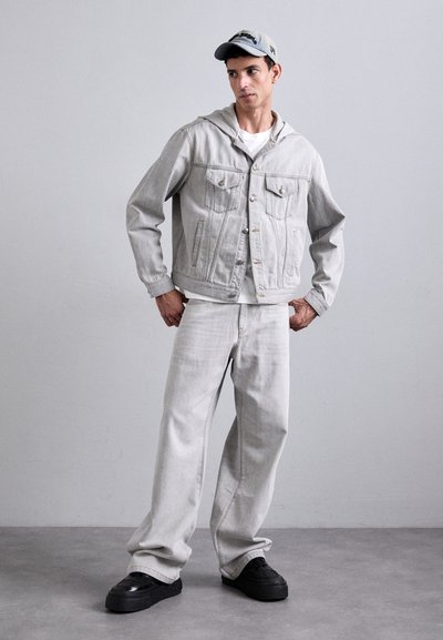 Veste en jean grise avec capuche, dotée de deux poches poitrine, assortie à un jean gris ample et des baskets noires. Une casquette vient compléter la tenue.