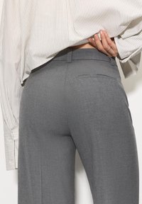 Szare dopasowane spodnie o gładkiej teksturze, z jedną tylną kieszenią i dopasowanym stanem. Model nosi koszulę w paski.