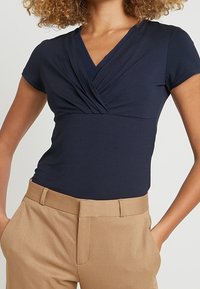 Marinblå kortärmad topp med V-ringning och rynkad framdel, matchad med beiga högmidjade byxor. Slät tygtextur.