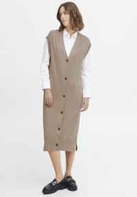 Vestito in maglia beige con scollo a V profondo, bottoni frontali, spacchi laterali e senza maniche, abbinato a scarpe con plateau nere.