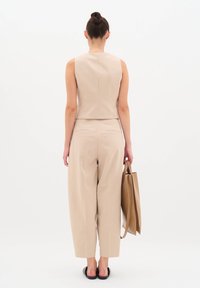 Beige bezrukávový top a kalhoty s vysokým pasem, hladká textura, minimalistický design. Drží hnědou tašku, stojí bos.