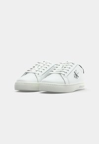 White leather sneakers with round toes, flat laces, a black heel accent, and a logo on the side. Textured rubber outsole for grip.