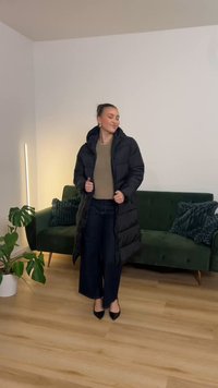 Černá prošívaná péřová bunda s kapucí. Nositelná přes béžové tričko a tmavé džíny. V pozadí zelená pohovka a pokojová rostlina.