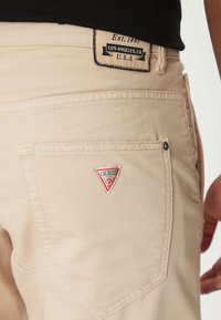 Jeans beige de Guess que presentan un bolsillo trasero con un logo triangular rojo y blanco y detalles de costura. La etiqueta dice "Los Ángeles, CA."