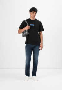 Mees, kes kannab musta Tommy Jeans T-särki, tumejuhuseid teksaseid, valgeid spordijalgu ja kellel on seljas väike hall õlakott, seistes ühtlasel valgel taustal.