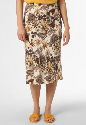 Femme portant une jupe crème longueur genoux avec des imprimés floraux tropicaux marron et jaune, et des sandales tressées jaunes à bouts ouverts, debout les bras détendus.
