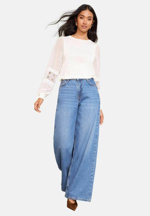 Fehér hosszú ujjú felső csipkével és pöttyös áttetsző ujjakkal, magas derekú, széles szárú kék farmernadrággal párosítva. Barna magassarkú cipő.