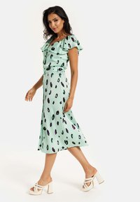 Mint green dress with frilled sleeves, loose fit, and black and purple spotted pattern. Paired with cream block-heeled sandals.